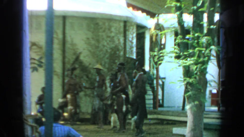 1964 Flushing Meadows frame showing performers in conical hats near a courtyard wall, watched from a partly obstructed visitor viewpoint.