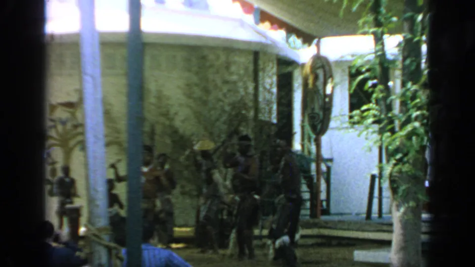 1964 Flushing Meadows home-movie frame of performers and spectators in a tropical-styled courtyard, partly obscured by dark foreground posts.