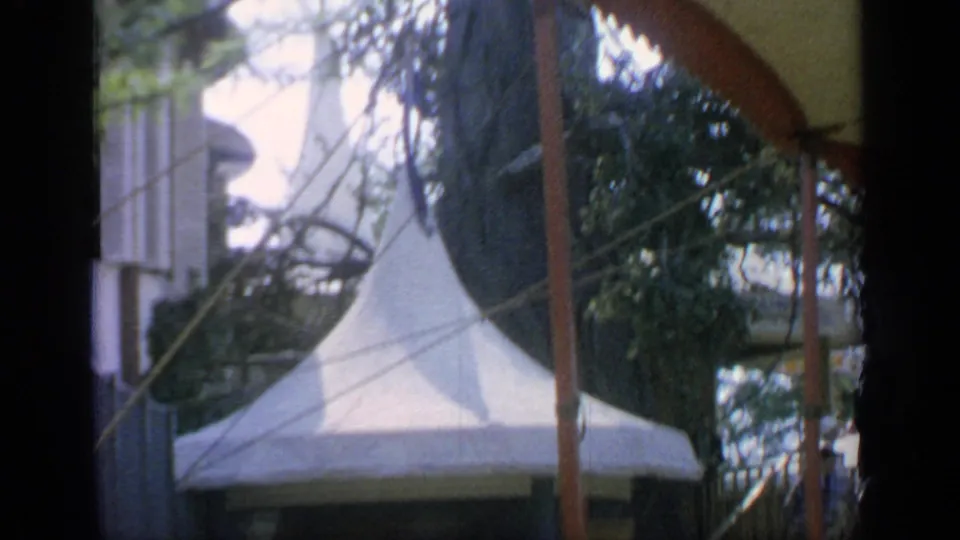 Visitor-shot 8mm view at Flushing Meadows with a white pointed tent, branches, poles, and fairground structures.