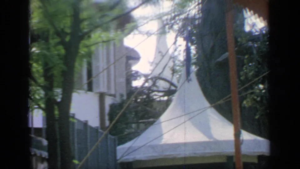 1964 Flushing Meadows fairground view with a pointed white tent, tree canopy, poles, and partially visible structures.
