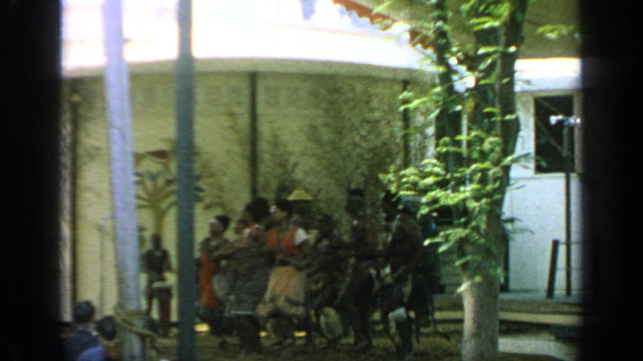 A group of dancers stands before spectators beside trees and columns.