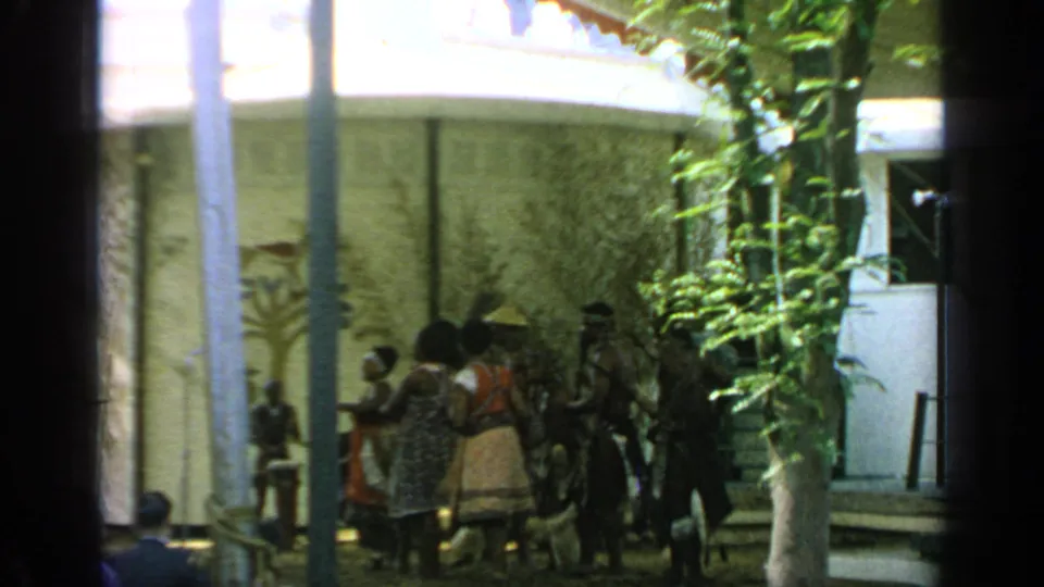 Visitor-shot 8mm frame of dancers in patterned garments moving near a small audience at the 1964 Flushing Meadows World's Fair.