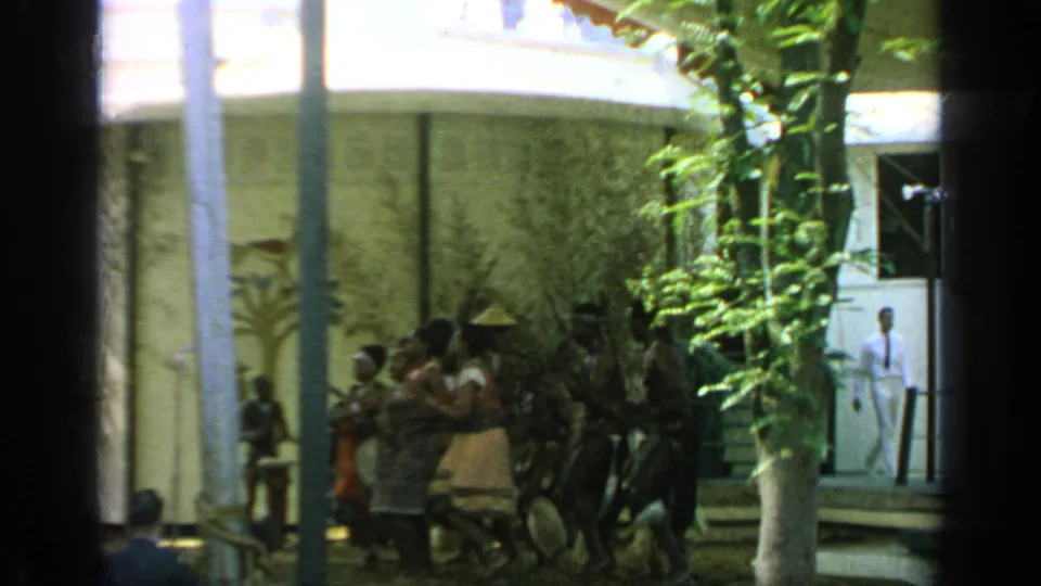 The performance continues in the Flushing Meadows courtyard while spectators watch and a person in white stands near a doorway.