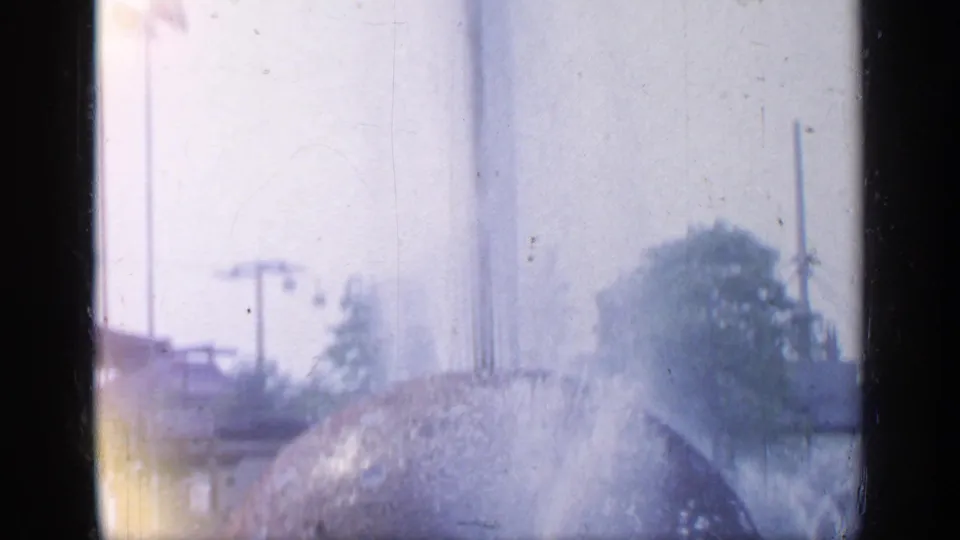 Faded 8mm view at Flushing Meadows showing fountain water spraying near a large spherical feature and distant outdoor structures.
