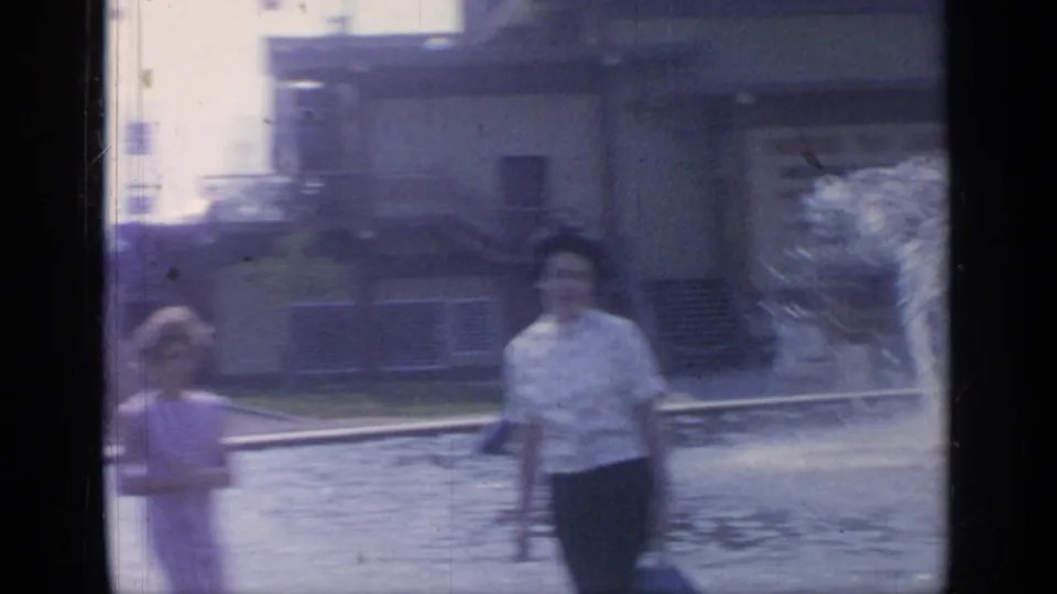 Flushing Meadows home-movie frame showing visitors near the fountain pool, including a child-sized figure and an adult in a light shirt.
