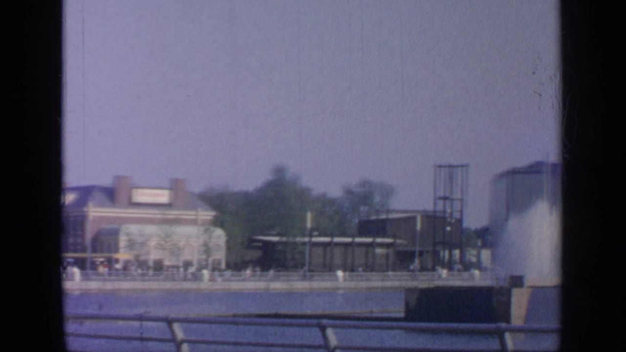 Waterfront fairground view with fountain, railings, trees, and modernist buildings.