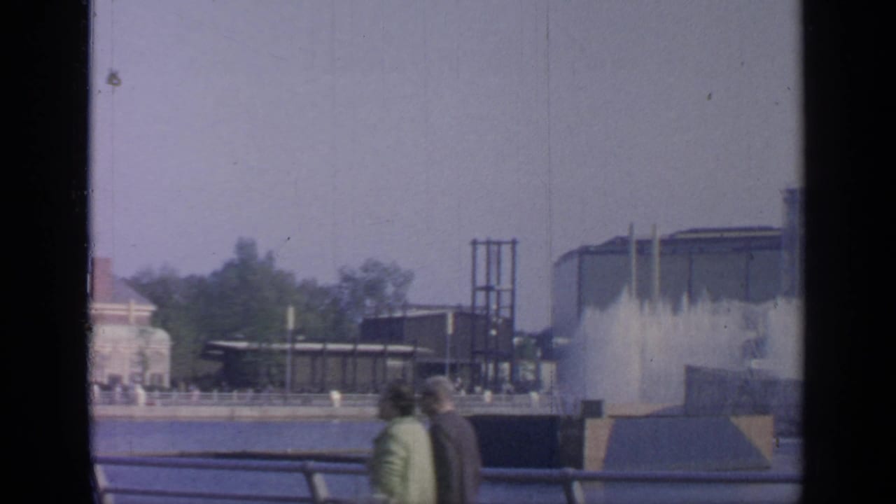 Two adults walk hand in hand along a Flushing Meadows promenade, with fountain spray and World's Fair-era structures in the background.
