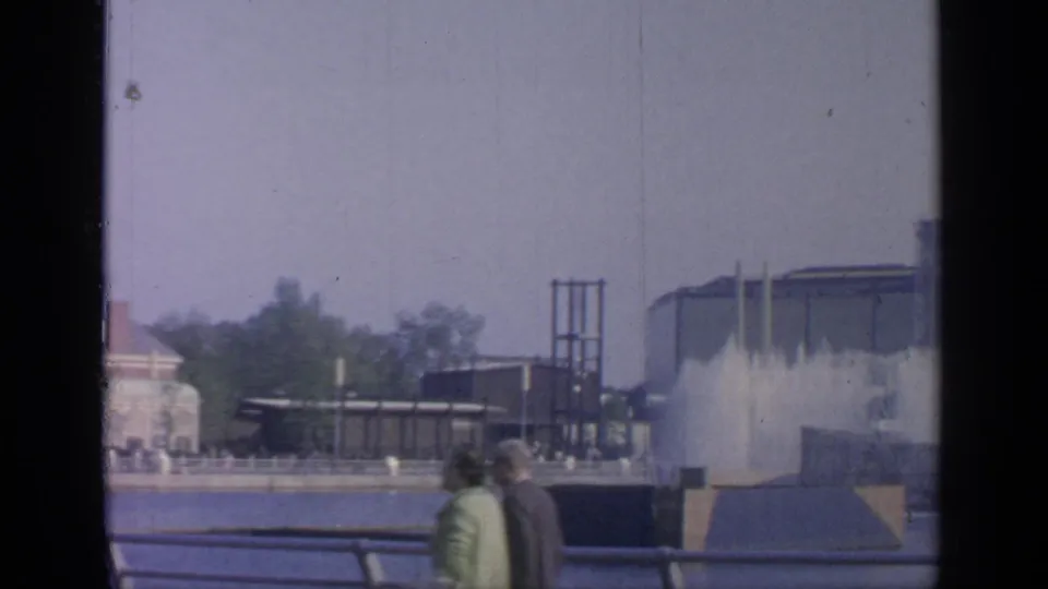 Two adults walk hand in hand along a Flushing Meadows promenade, with fountain spray and World's Fair-era structures in the background.
