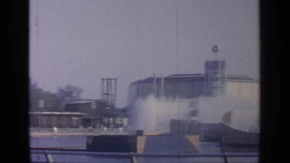 Flushing Meadows fairground architecture and fountain spray fill the frame, seen from across a waterfront railing on 8mm film.