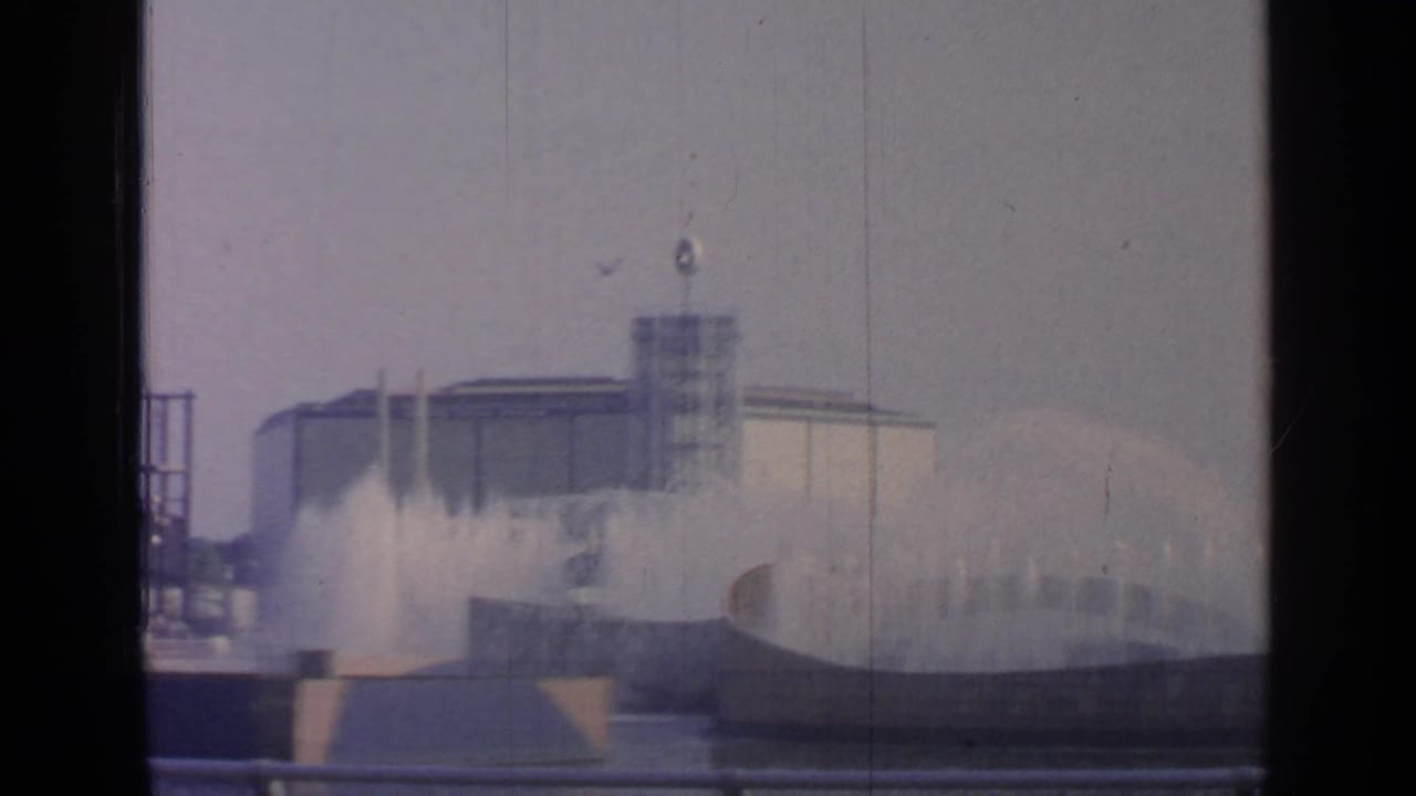 Fountain jets rise in front of a modern building and tower behind railings.