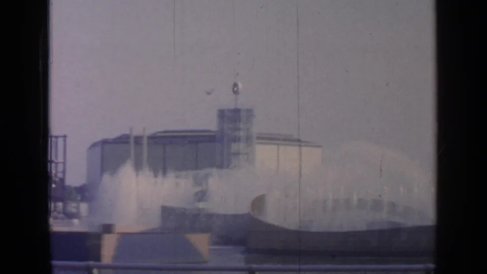 1964 Flushing Meadows fountain display with water spray, a towered structure, overcast sky, and dark 8mm frame edges.