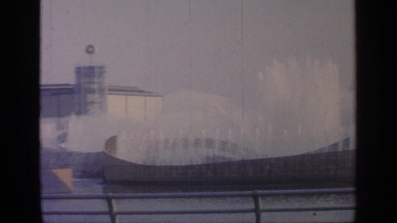 High water spray at Flushing Meadows in 1964, seen across railings with a towered fairground structure in the background.