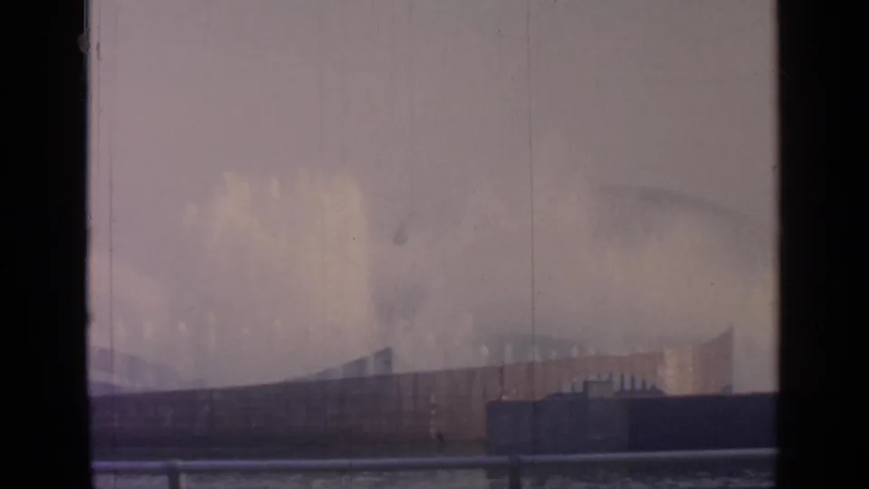 Wide spray plume at Flushing Meadows in 1964, with railings, low structures, haze, and visible 8mm film borders.