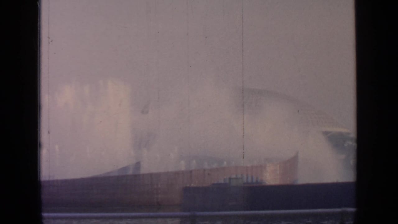 Distant fairground fountains seen through dark window framing with a hazy structure behind them.