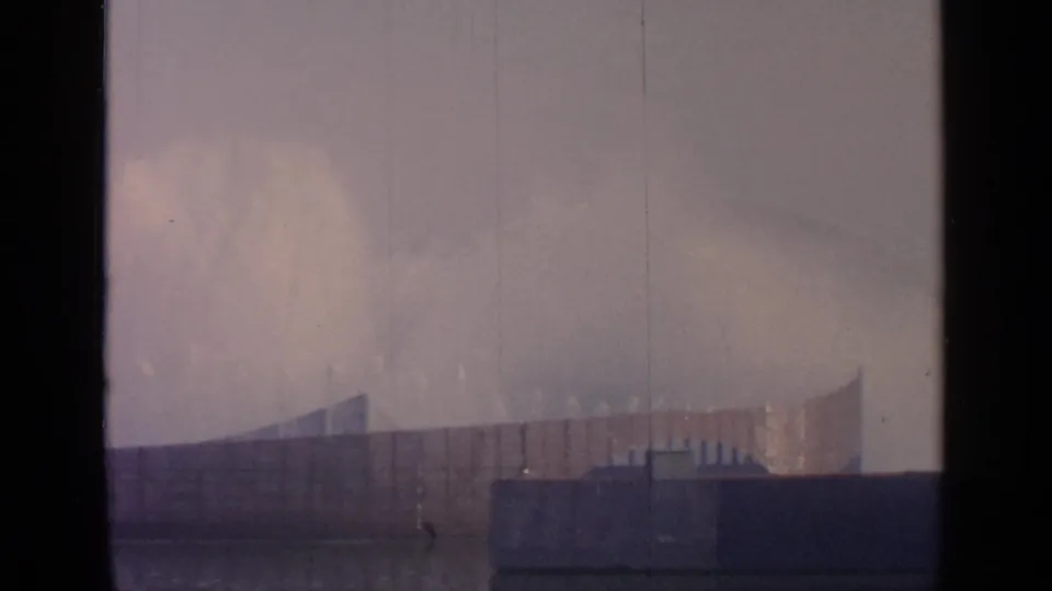 The final 1964 Flushing Meadows view holds on misting fountains, low fair buildings, and hazy sky seen through a dark interior window edge.
