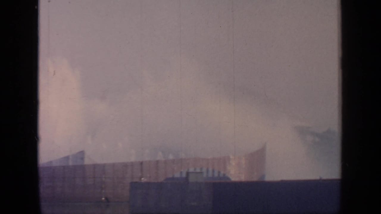 Hazy fountain spray beyond a dark foreground barrier with distant fairground structures.