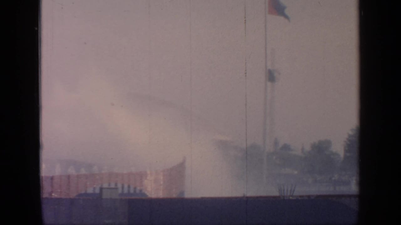 Flagpole and distant fountain spray seen through haze with trees at the right edge.