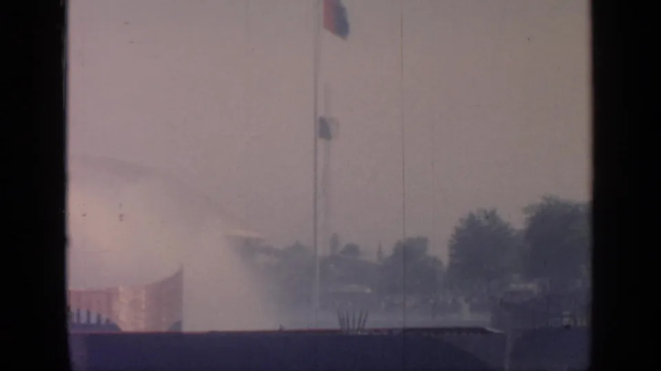 Hazy 1964 Flushing Meadows view with flagpole, fountain spray, trees, and dark foreground barrier.