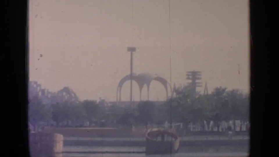 Hazy 8mm view of Flushing Meadows with water in front, trees, arched fair structures, and a floodlight tower beyond.