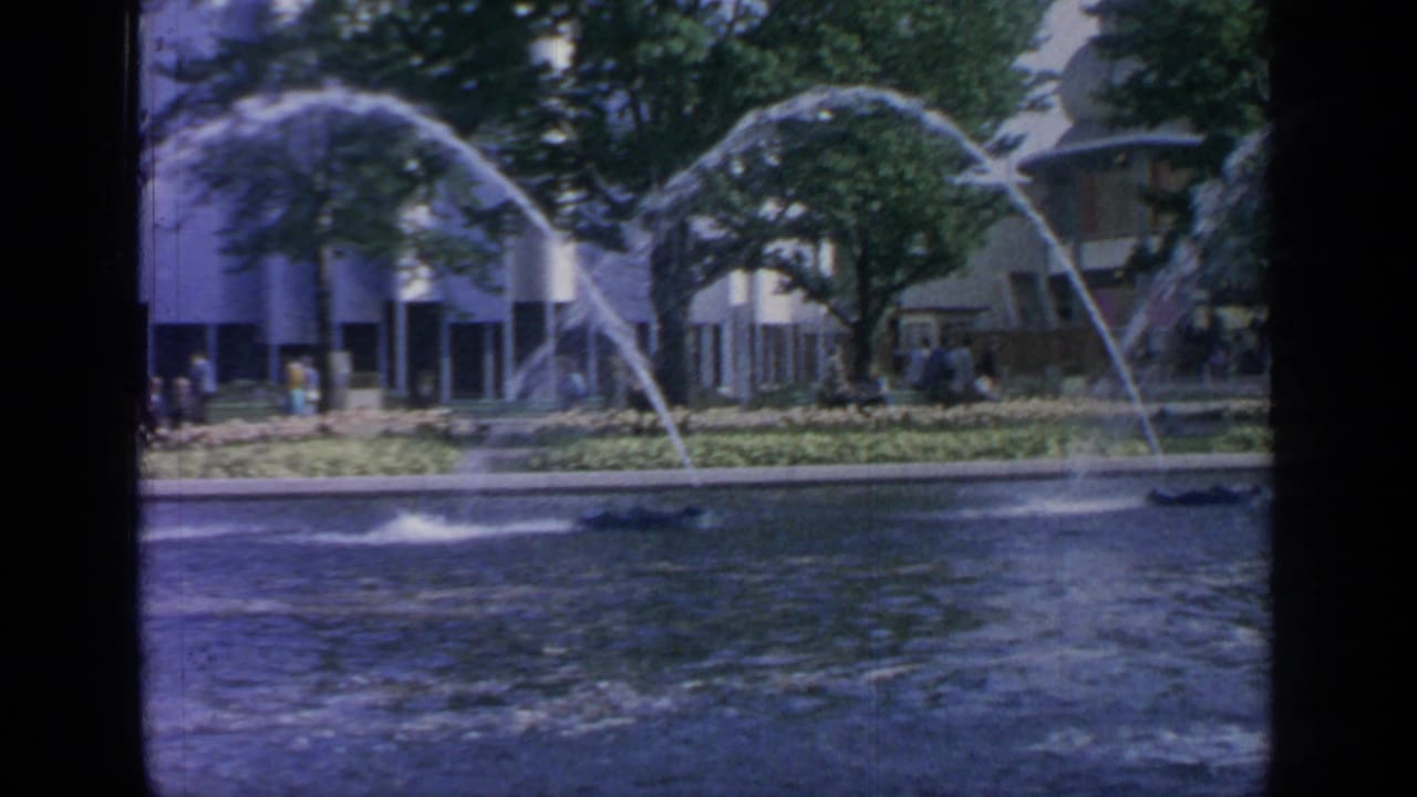 Large pond with arcing fountain jets, trees, visitors, and modern buildings beyond.
