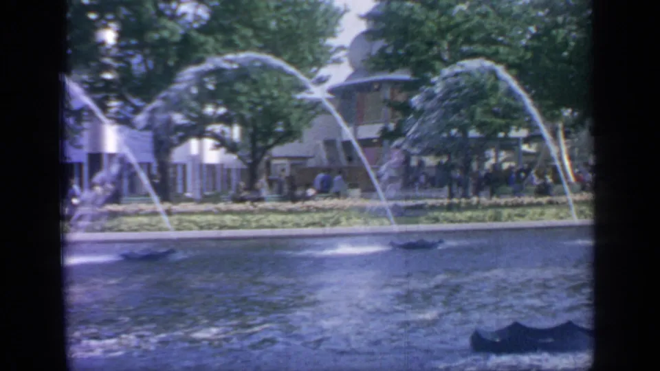 1964 Flushing Meadows fountain scene with multiple water arcs over a pond, trees, exhibition buildings, and small visitor groups along the path.
