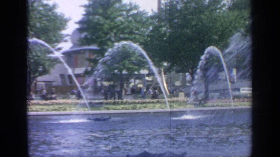Flushing Meadows pond view showing fountain jets, visitors on the far path, trees, and a rounded mid-century building in the background.