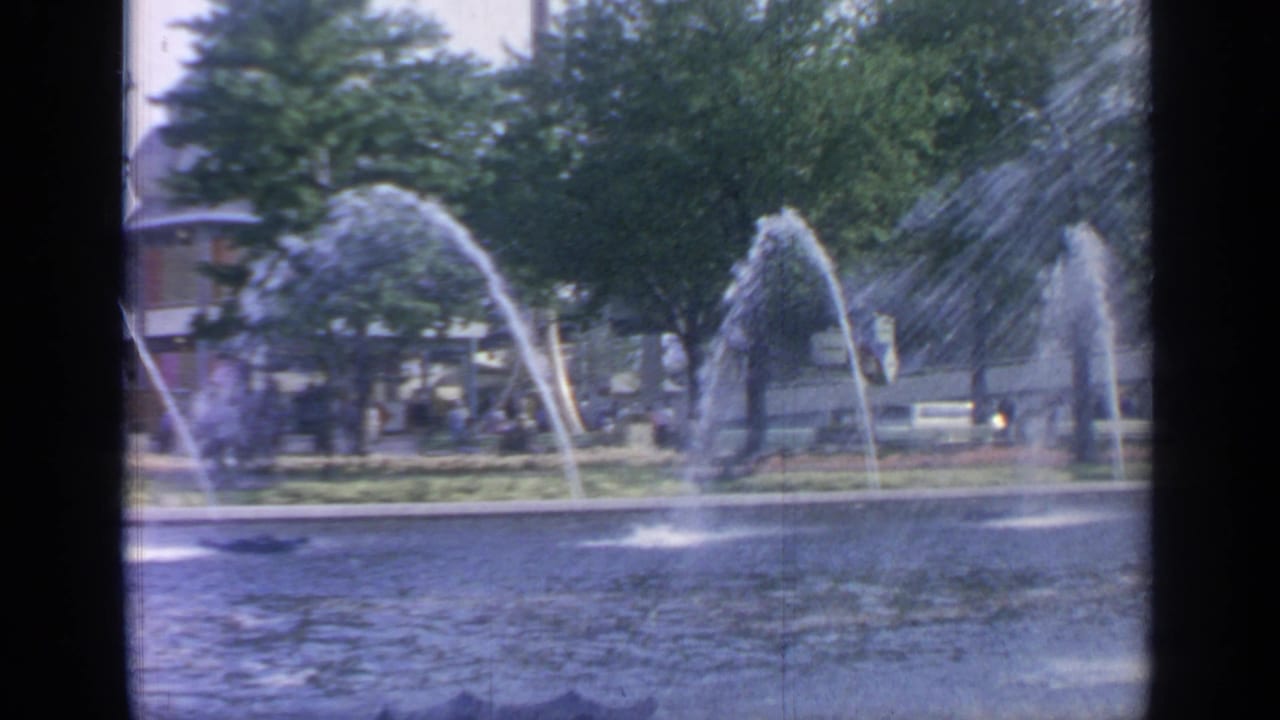 Arcing fountain jets over a large pool with trees and distant visitors behind them.
