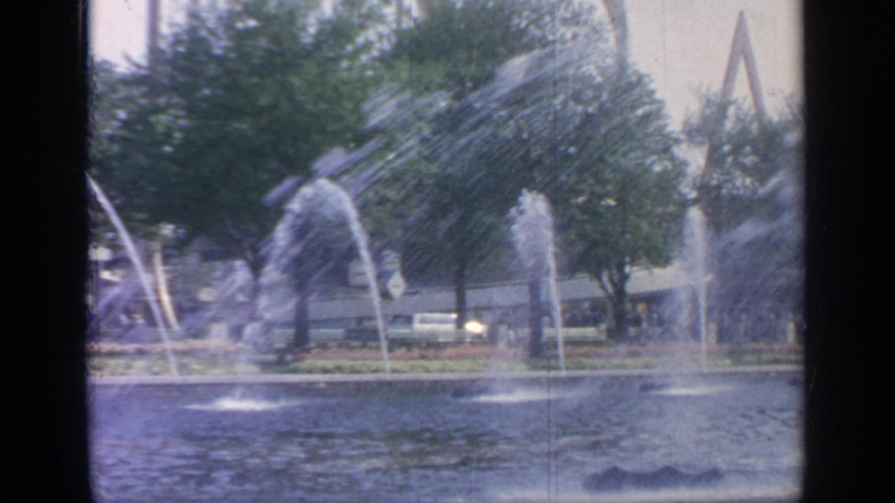 Water jets spray across the Flushing Meadows World's Fair pool, with landscaped grounds and angular structures visible behind trees.