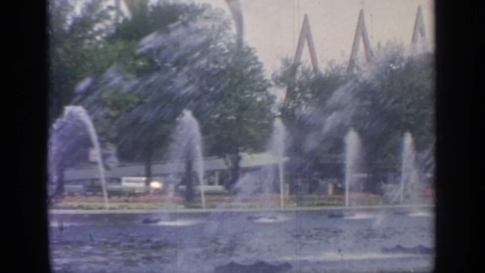 8mm view of the World's Fair fountain display, showing tall water jets, a broad pool, trees, benches, and angular fair structures.