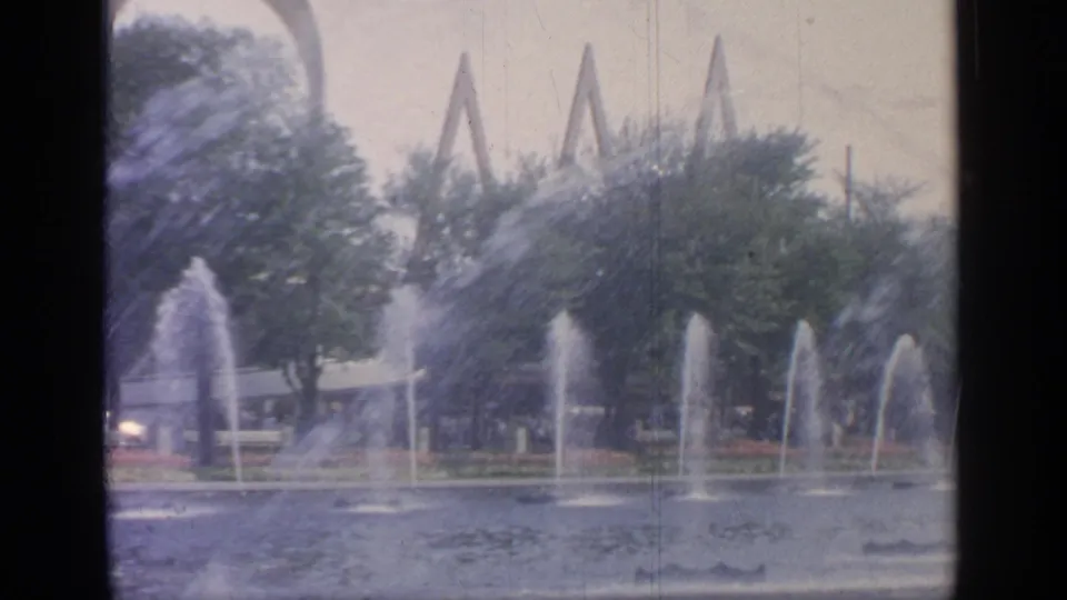 Wide 8mm frame of the Flushing Meadows fountain pool, with several jets, rippling water, trees, benches, and distant fair structures.