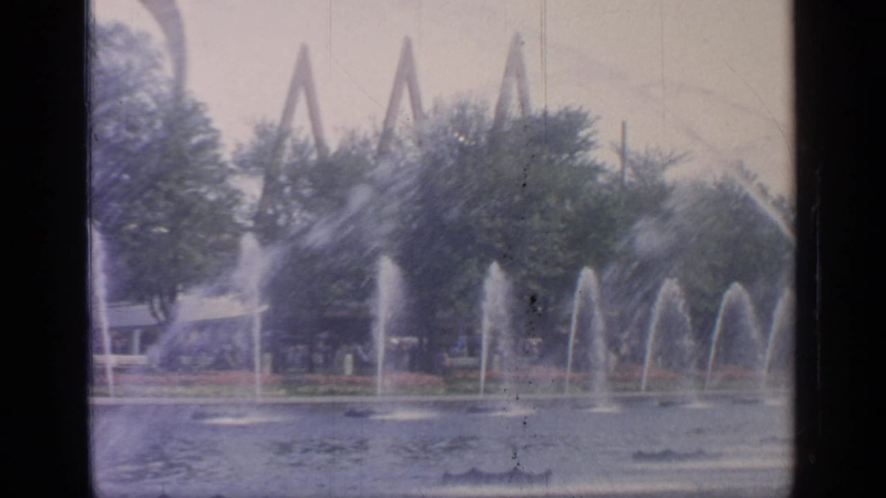 1964 Flushing Meadows fairground view with water spouts crossing a pond and angular structures partly visible behind trees.