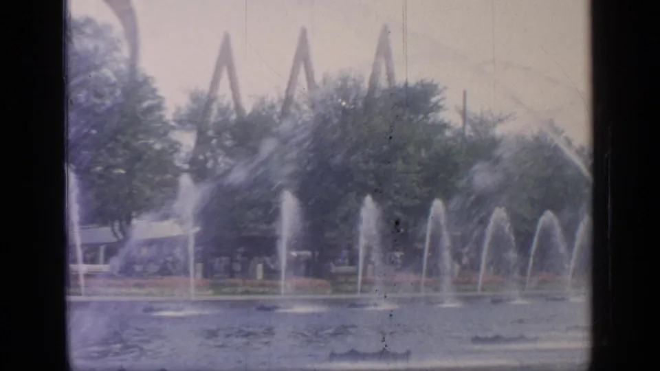 1964 Flushing Meadows fairground view with water spouts crossing a pond and angular structures partly visible behind trees.