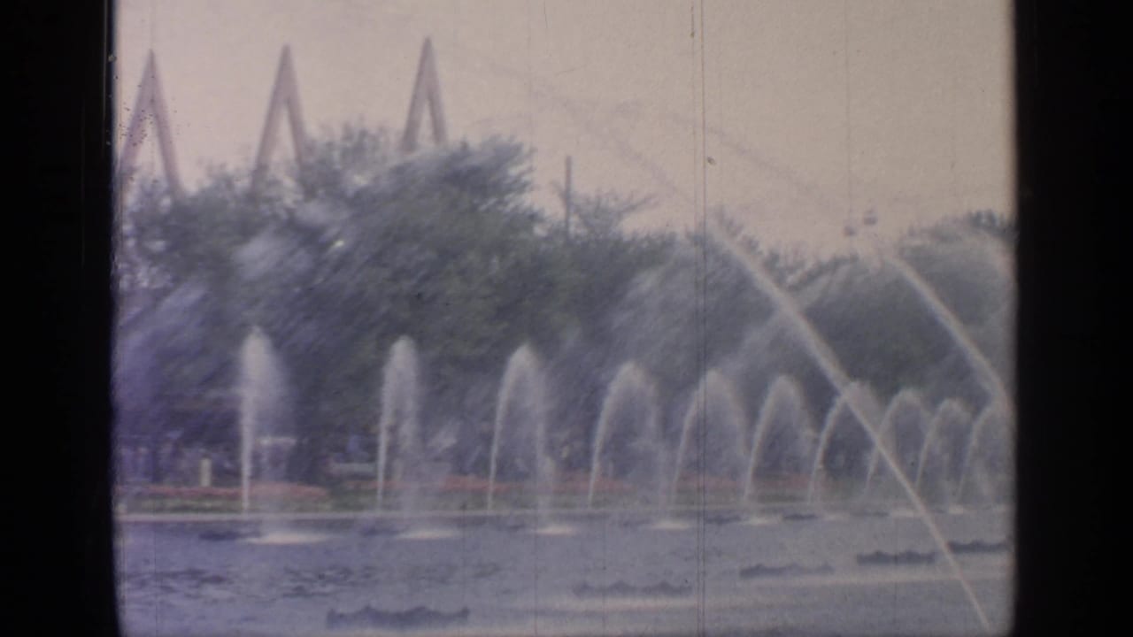 Arcing fountain streams cross a pond with trees and triangular structures behind them.