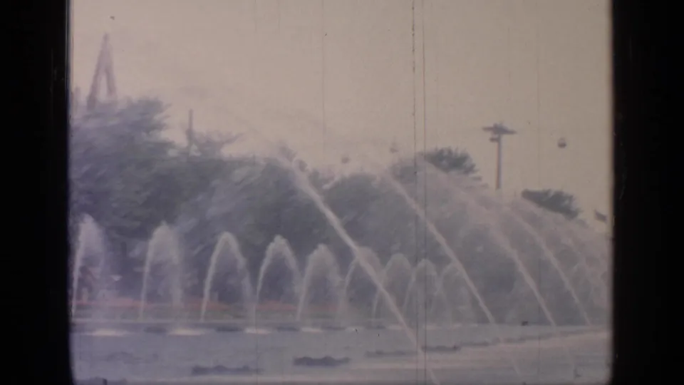 1964 World's Fair fountain display seen from across the pond, with layered water jets and a fairground light pole behind the trees.