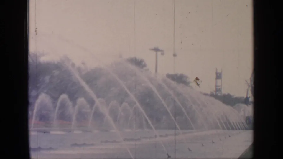 Flushing Meadows fountain scene with repeated water arcs across the pond and distant fairground structures softened by 8mm film texture.