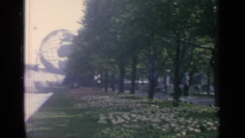 A visitor's 8mm view looks along trees and flower beds toward the Unisphere and fountain area.
