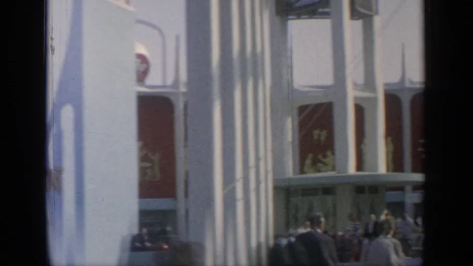 Handheld 8mm view of white World's Fair pavilion columns and red graphic panels, with visitors gathered along the lower edge.