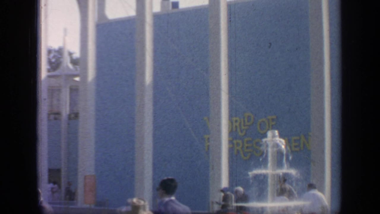 1964 fairground view with a blue pavilion facade, partial yellow sign lettering, fountain spray, and visitors.