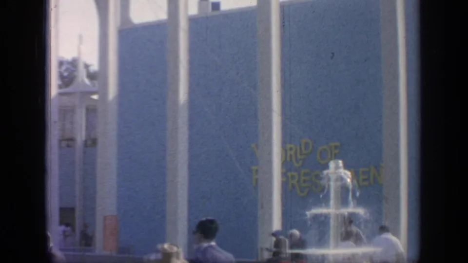 1964 fairground view with a blue pavilion facade, partial yellow sign lettering, fountain spray, and visitors.