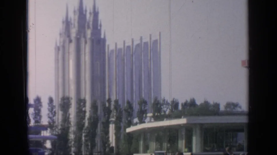 A distant spired World's Fair structure rises beyond trees and a low curved building in Flushing Meadows.