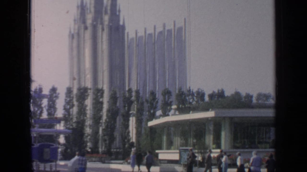 Visitors walk near a low building below tall fairground spires.