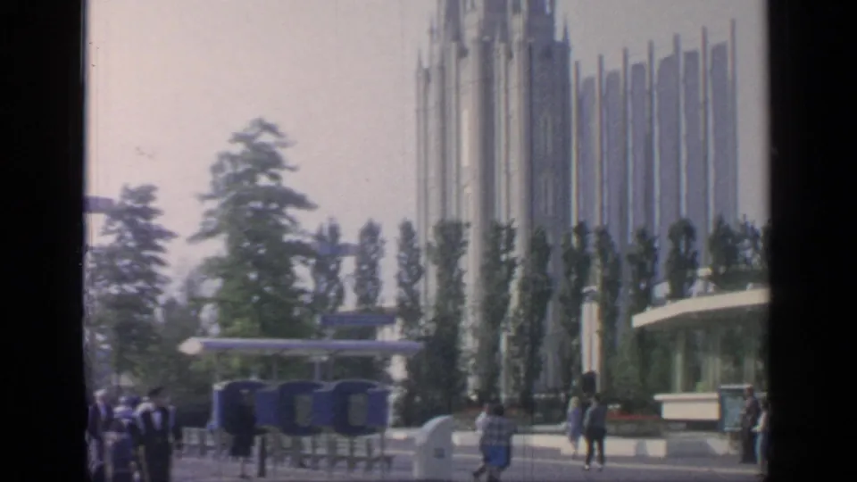 A visitor-shot view of Flushing Meadows shows people crossing the grounds near blue fairground fixtures, trees, and pavilion spires.