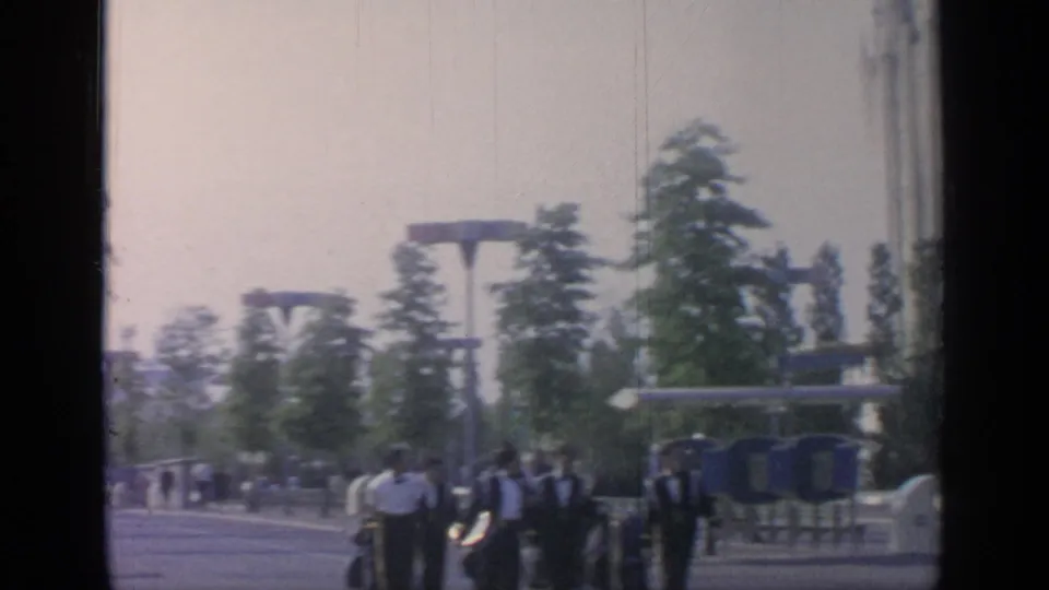 At Flushing Meadows, several visitors, including men in white shirts and dark bow ties, gather on a broad walkway near trees and fair fixtures.