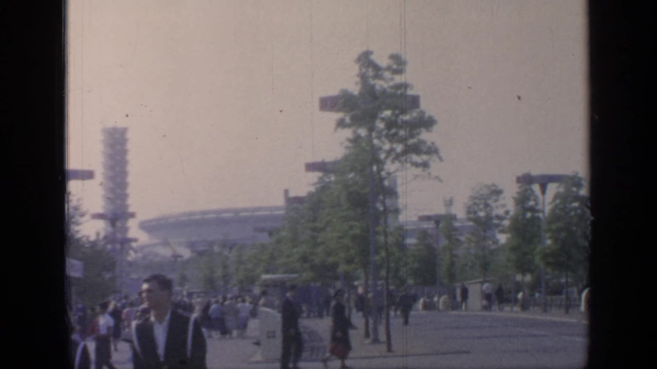 Crowds on a fairground walkway with trees, light poles, and a distant round structure.