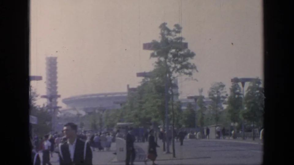 Visitors at Flushing Meadows in 1964 walk past trees and fairground lighting, with a distant round modern structure in view.