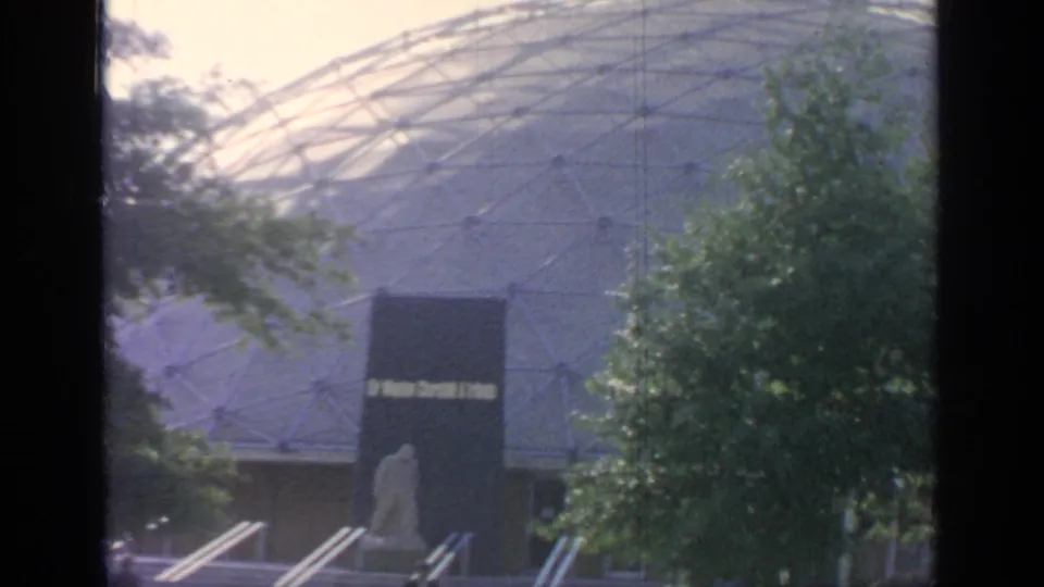 White geodesic-style dome at Flushing Meadows, partly obscured by trees, with a dark entrance area below.