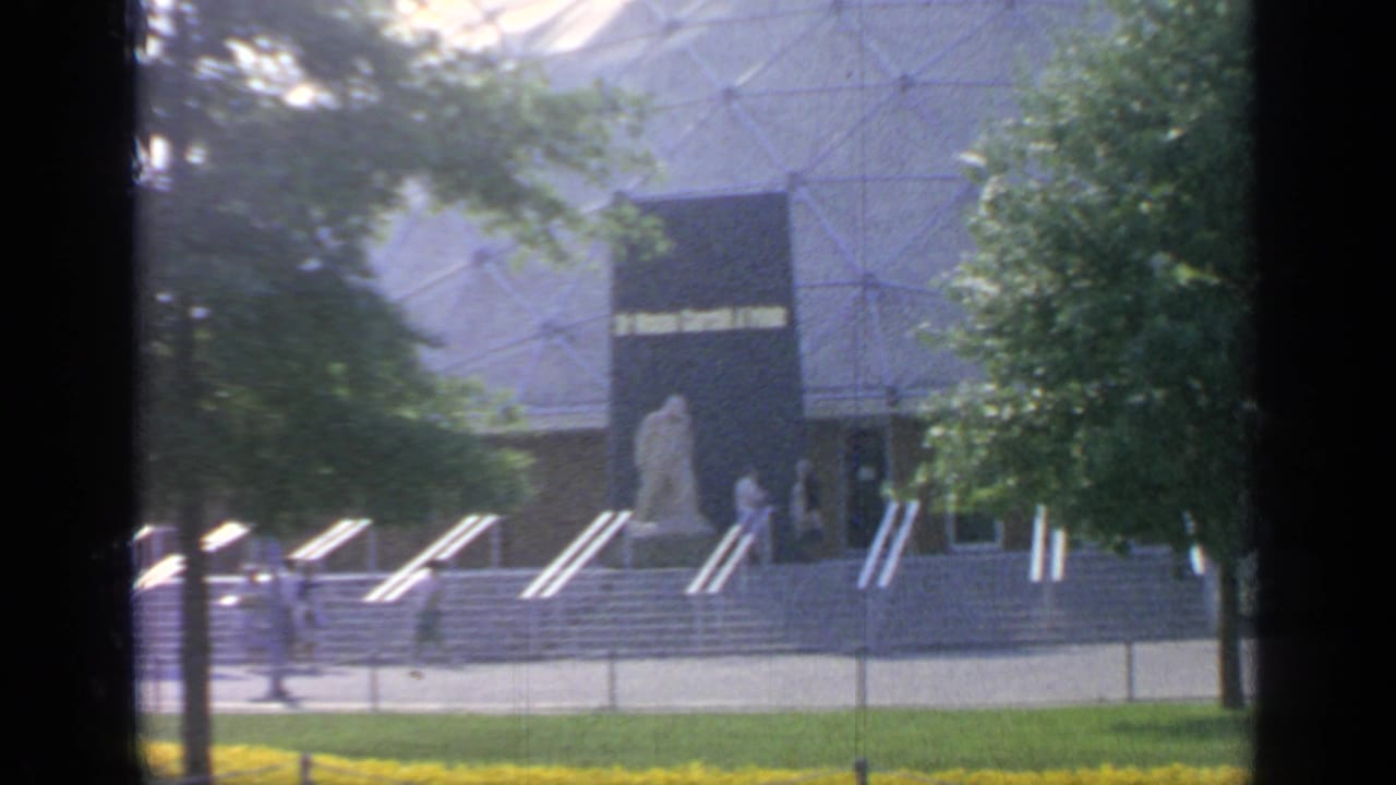 Visitors walk near flower beds and steps in front of a large fairground building.