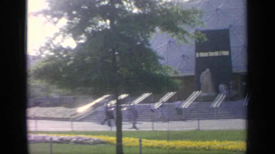 At Flushing Meadows in 1964, visitors cross the fairgrounds near trees, yellow flowers, steps, and a large modern building.