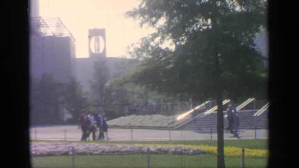 1964 Flushing Meadows home-movie frame with visitors walking past white and yellow flowers below fairground steps and towers.
