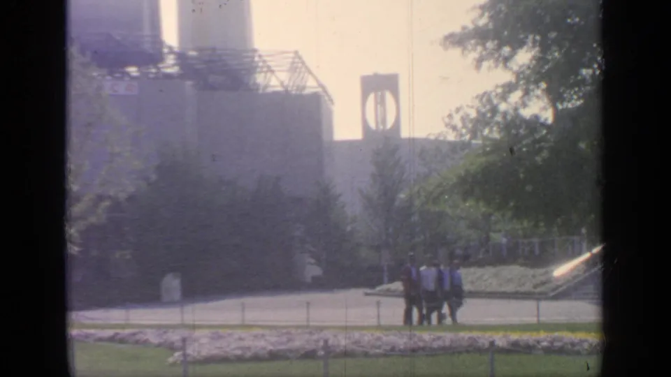 A 1964 Flushing Meadows frame shows visitors passing flower beds, with fairground steps, trees, and a tower-like structure behind them.